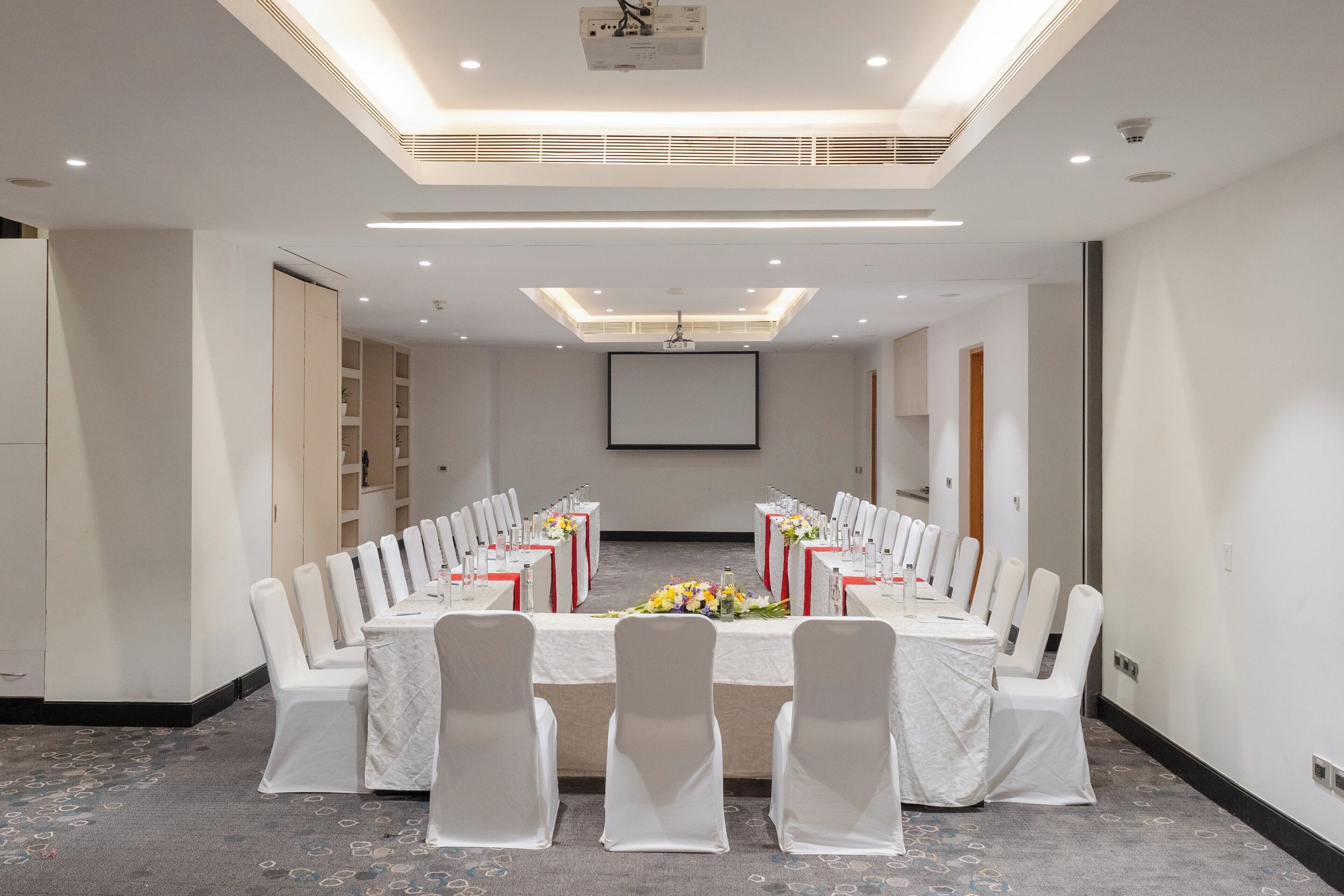 A conference room with a long table, white chairs, and a projector screen.