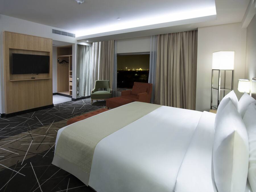 A hotel room with a large bed, a TV mounted on the wall, a seating area with chairs, and a view of the city at night.