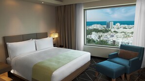 Habitación de hotel con una cama grande, una silla, una mesita de noche, una lámpara y vistas de un paisaje urbano a través de un ventanal amplio.