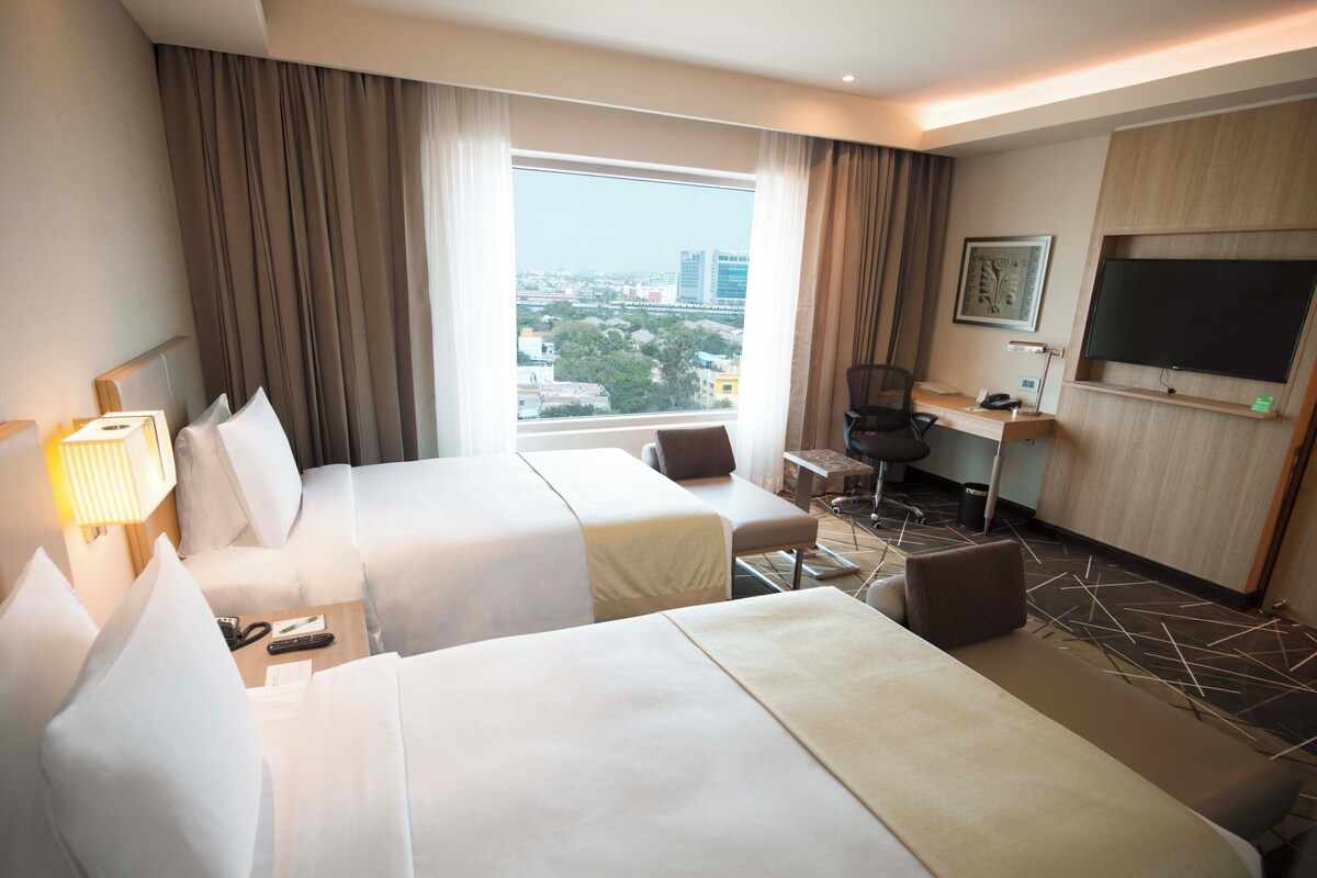 Une chambre d’hôtel avec deux lits, un bureau, une chaise, une télévision et une fenêtre donnant sur la ville.