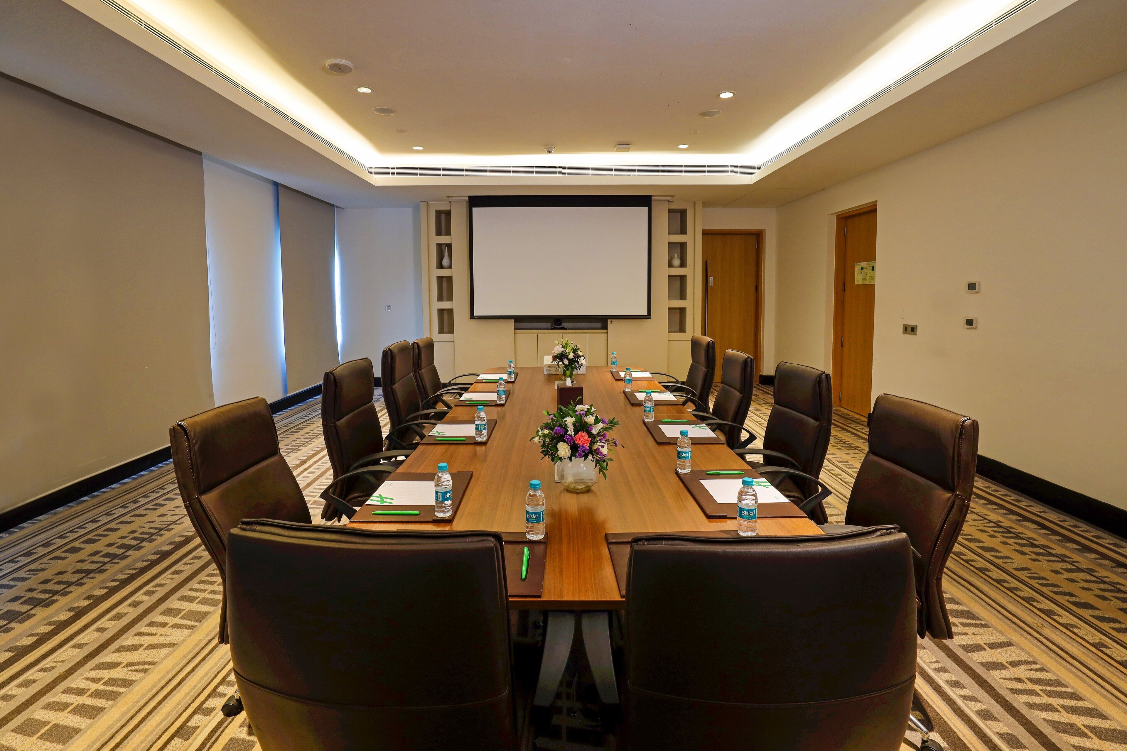 A conference room with a long table, chairs, a projector screen, and a floral centerpiece.