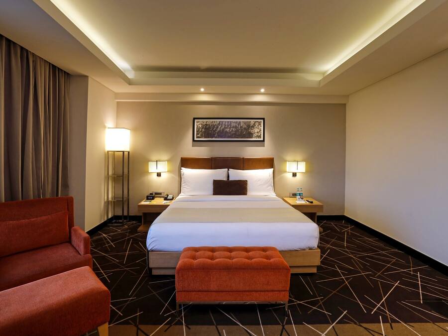 A hotel room with a bed, two bedside lamps, a red armchair, a red ottoman, and a framed picture on the wall.
