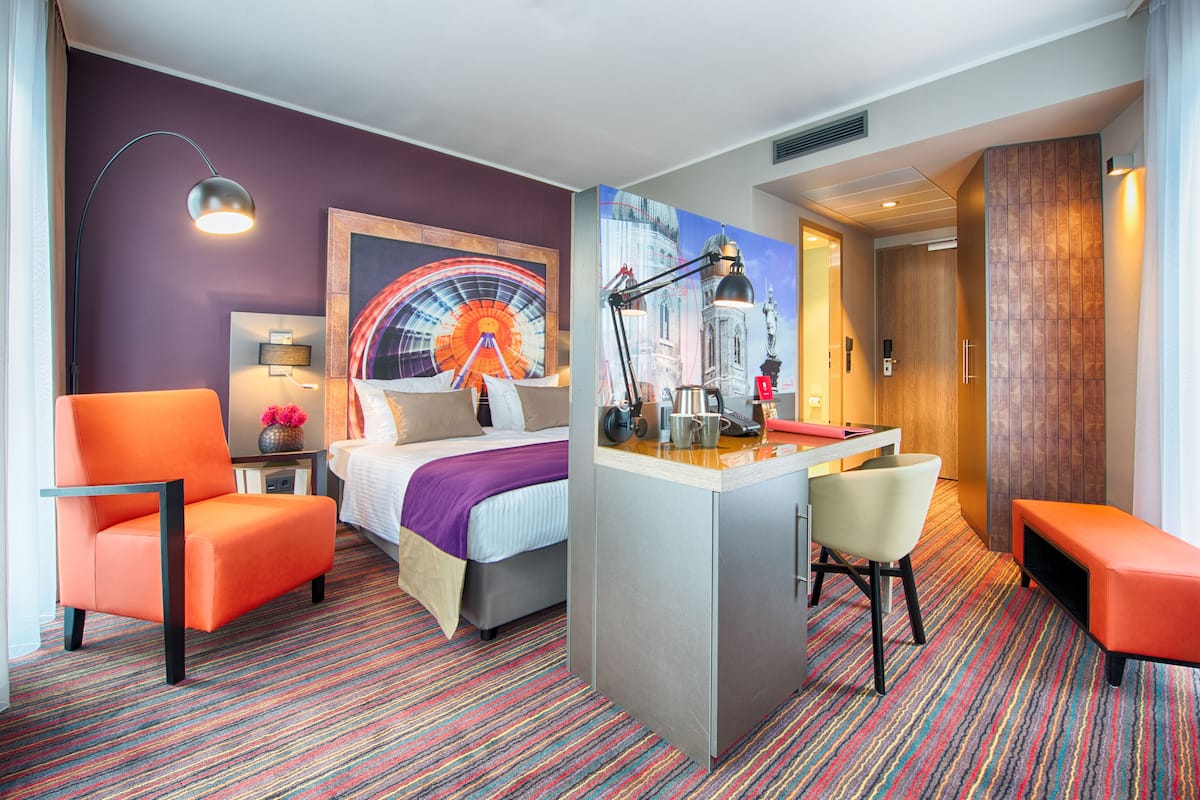 A hotel room with a bed, a desk, a chair, a bench, a lamp, and a colorful carpet.