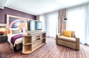 A hotel room with a bed, a TV, a sofa, and a large window with curtains. - Leonardo Hotel Munich City South (Munich)