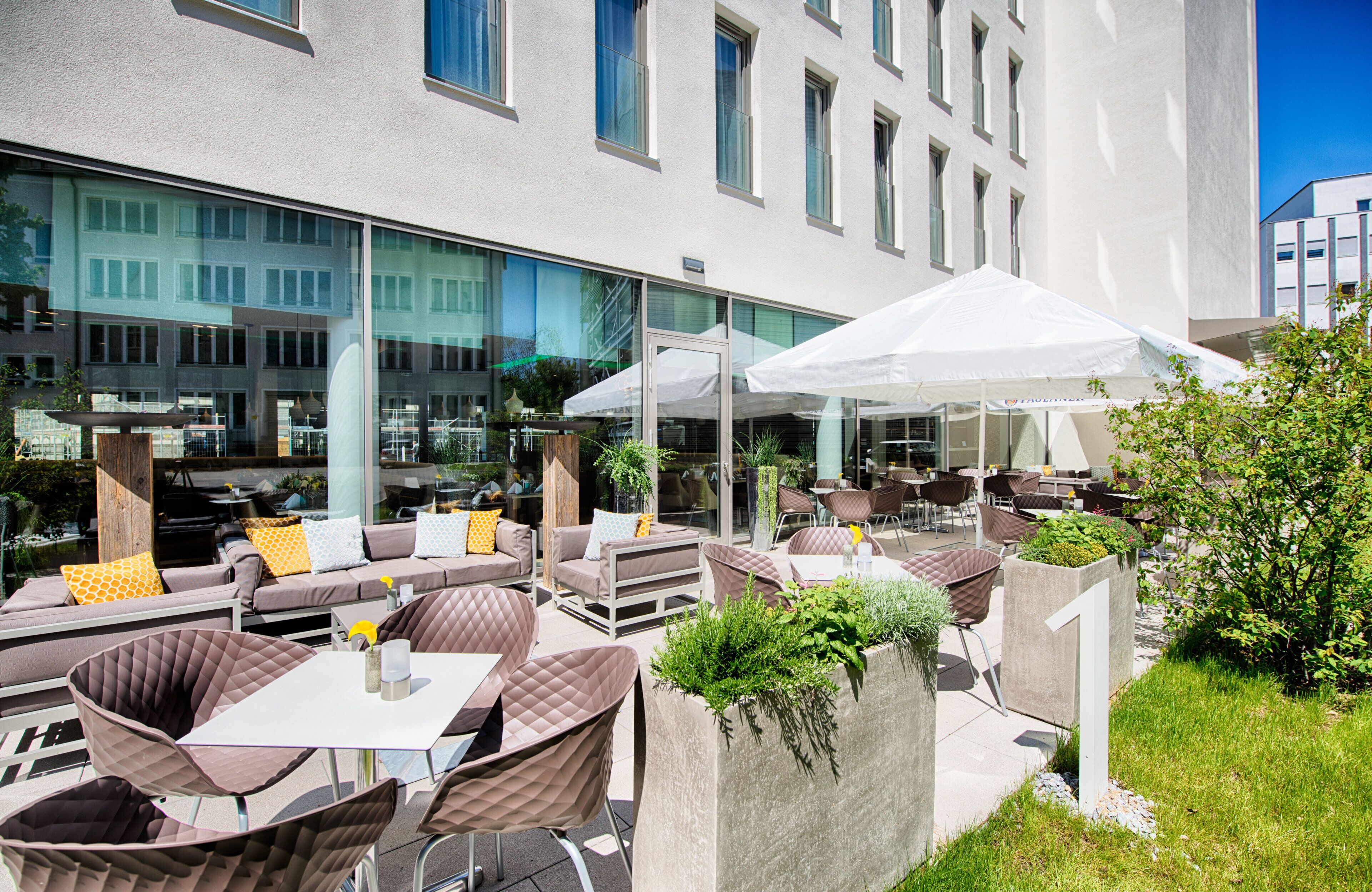 An outdoor seating area with tables and chairs, a white umbrella, and a building with large windows in the background.