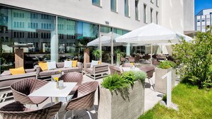 An outdoor seating area with tables and chairs, a white umbrella, and a building with large windows in the background. - Leonardo Hotel Munich City South (Munich)