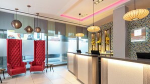 A hotel reception area with a modern design, featuring a reception desk, two red upholstered chairs, and decorative wall panels. - Leonardo Hotel Munich City South (Munich)