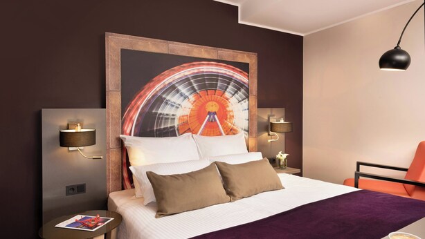 A hotel room with a bed, a nightstand, a reading lamp, a wall-mounted TV, a coffee table, and a chair. - Leonardo Hotel Munich City South (Munich)