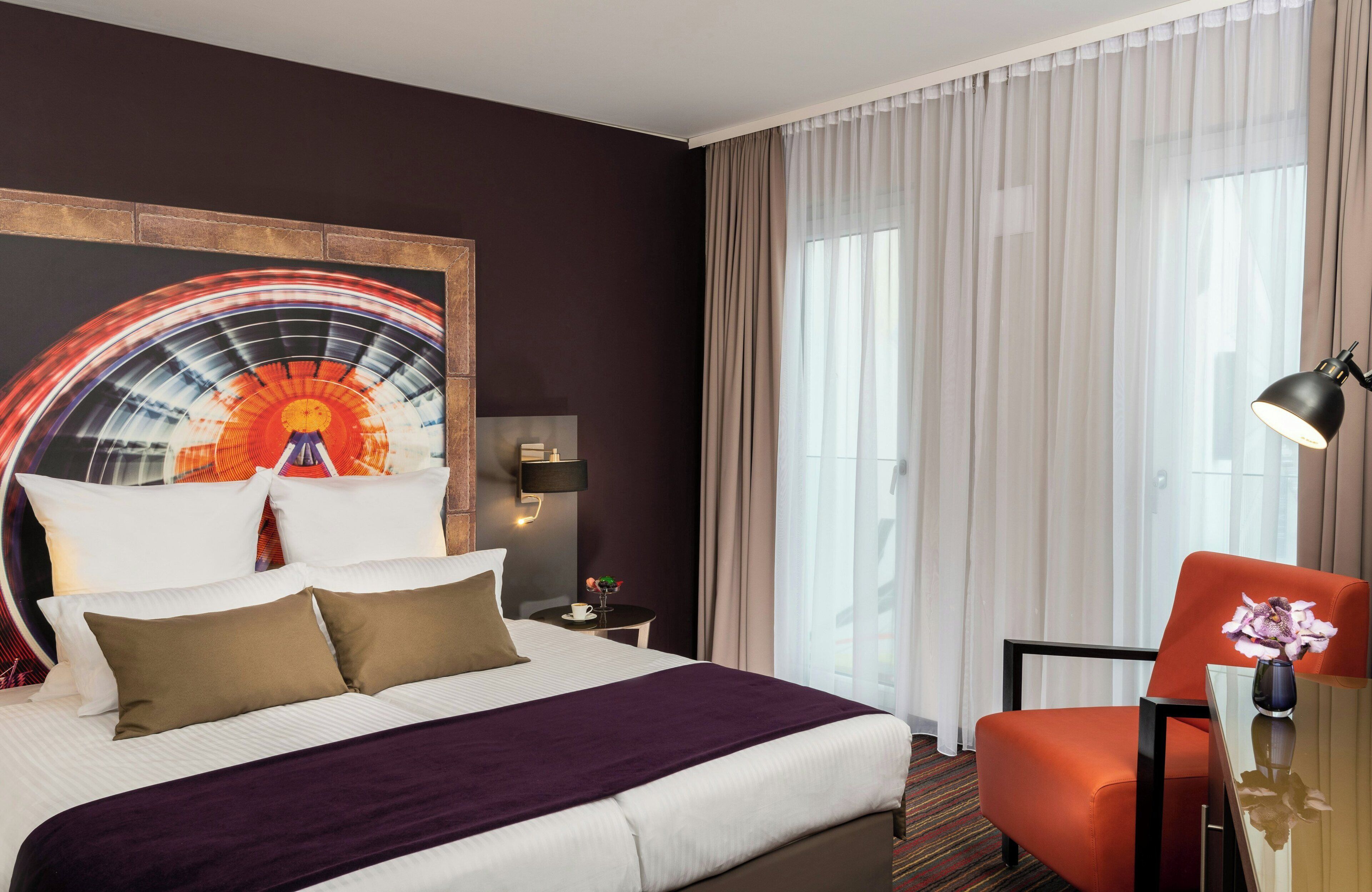Ein Hotelzimmer mit einem Bett, einem Schreibtisch mit Stuhl, einer Stehlampe, einer Wandleuchte, einem Fenster mit Vorhängen und einem Wandbild.