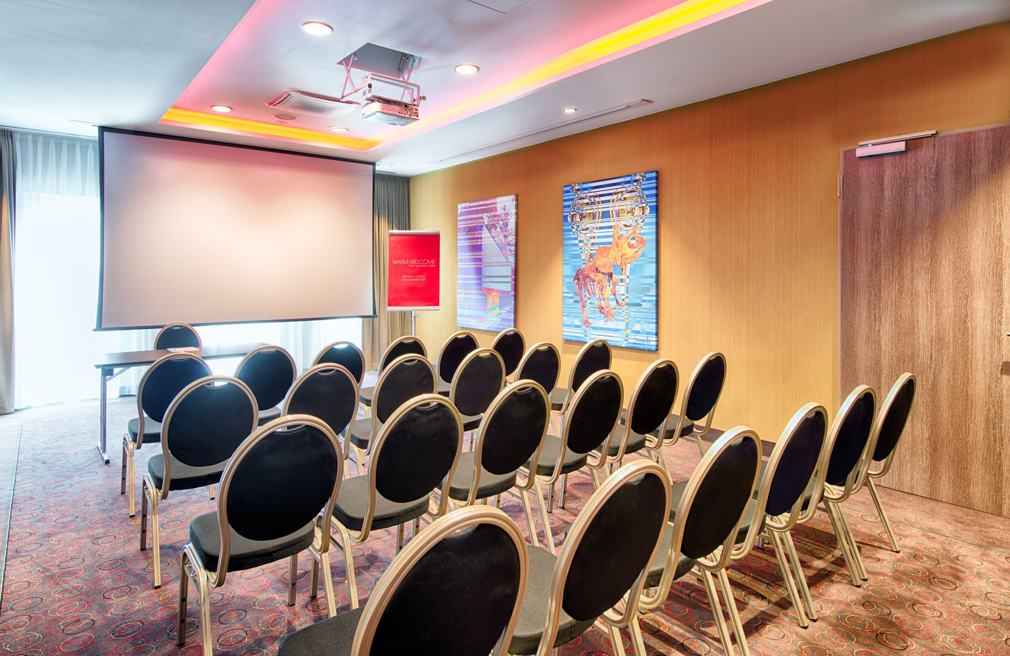 Un salón de conferencias con filas de sillas, una pantalla de proyector, un letrero de bienvenida rojo y dos grandes cuadros abstractos en la pared.