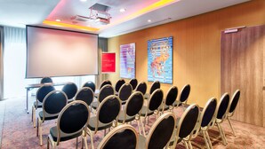 A conference room with rows of chairs, a projector screen, a red welcome sign, and two large abstract paintings on the wall. - Leonardo Hotel Munich City South (Munich)