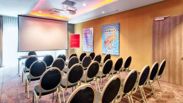 Un salón de conferencias con filas de sillas, una pantalla de proyector, un letrero de bienvenida rojo y dos grandes cuadros abstractos en la pared.