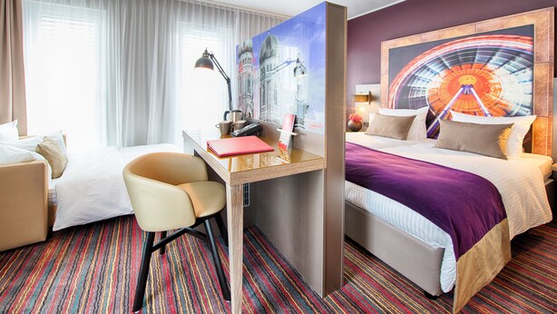 A hotel room with a bed, a desk with a chair, a window with curtains, and a large wall painting. - Leonardo Hotel Munich City South (Munich)