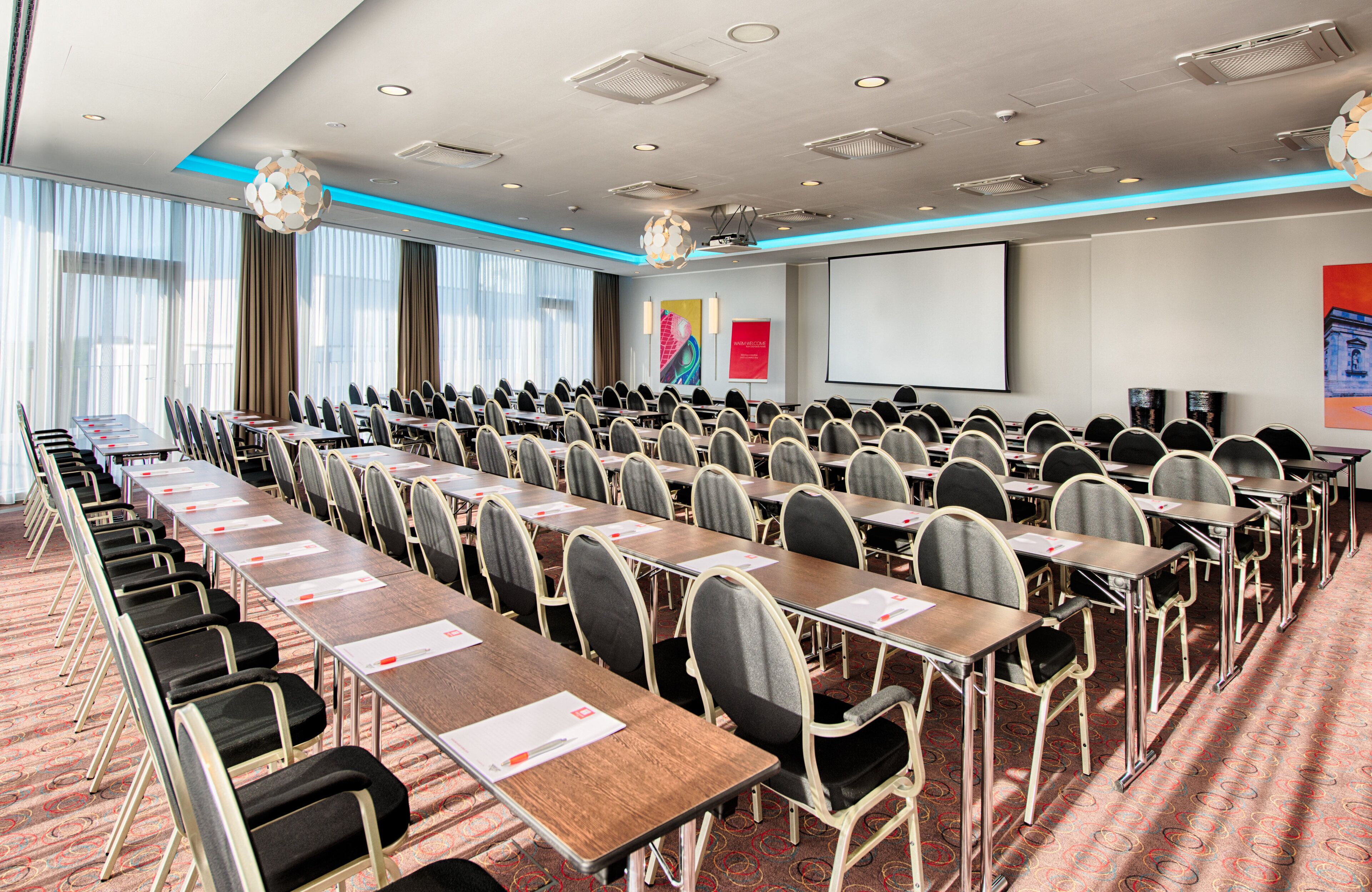A conference room with rows of tables and chairs, a projector screen, and a large window with curtains.