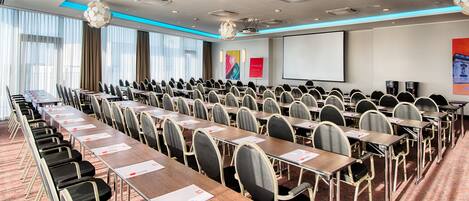A conference room with rows of tables and chairs, a projector screen, and a large window with curtains.