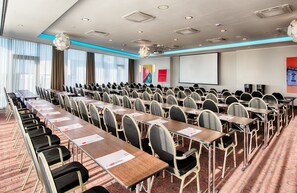 Un salón de conferencias con filas de mesas y sillas, una pantalla de proyector y un amplio ventanal con cortinas.