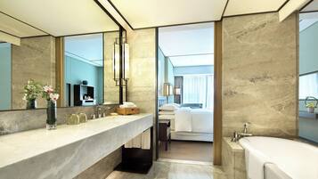 A modern bathroom with a marble countertop, a large mirror, a bathtub, and a view into the bedroom.
