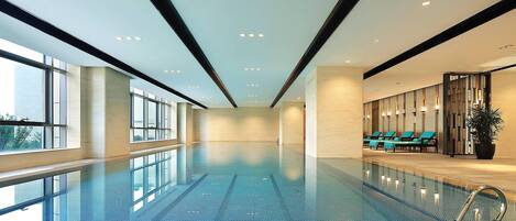 A large indoor swimming pool with clear blue water, surrounded by large windows and a seating area with lounge chairs.