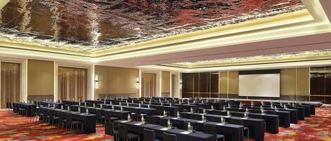 Un salón de conferencias con filas de mesas y sillas, una pantalla grande y un techo con superficie reflectante.