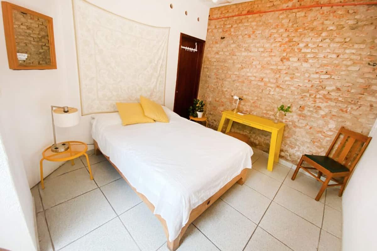 Un dormitorio con cama, mesita de noche, silla, lámpara y pared de ladrillo.
