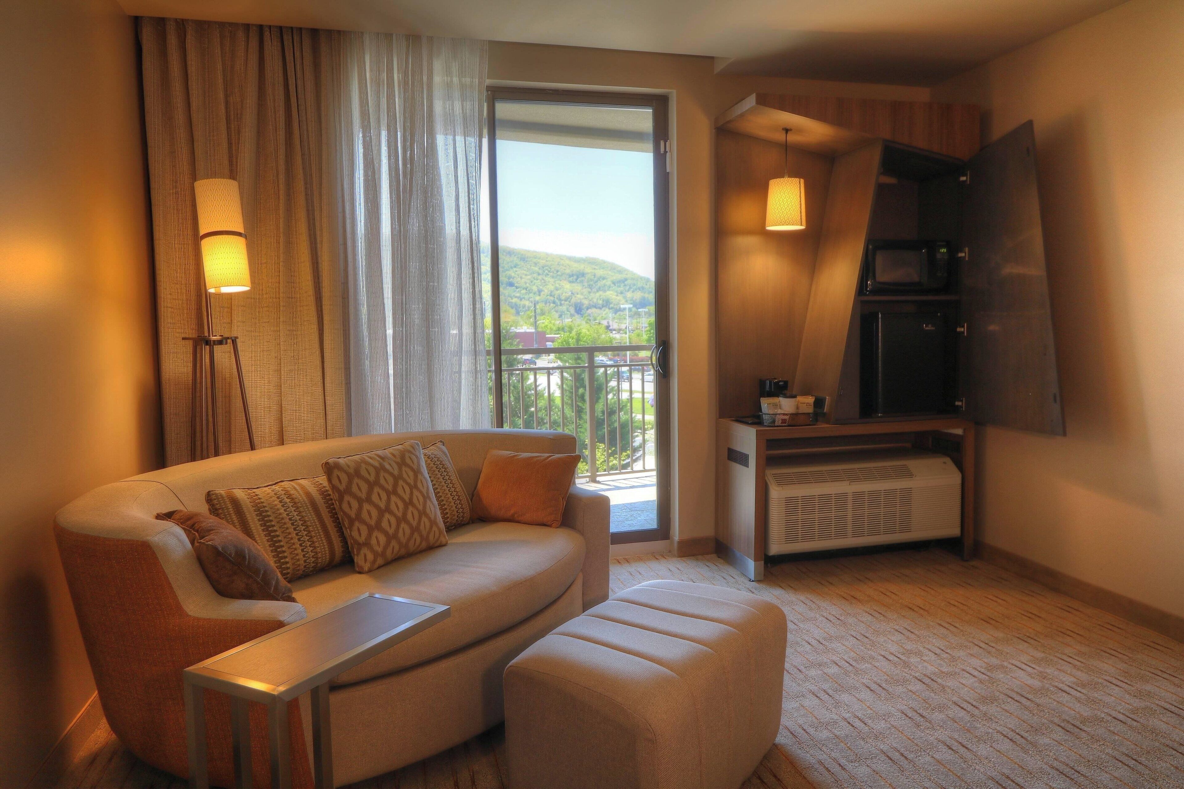 Una habitación de hotel moderna con sofá, reposapiés y una zona de comedor con microondas.