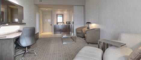 Quarto de hotel com escrivaninha, cadeira de escritório, sofá e uma pequena mesa. Há um espelho sobre a escrivaninha e uma luminária na mesa. O quarto possui piso coberto por carpete e vista para o corredor.