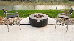 Un foyer circulaire entouré de deux chaises en métal, installé sur une terrasse en béton et offrant une vue sur une étendue d’eau.