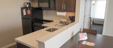 A compact kitchen with dark wooden cabinets, a stainless steel refrigerator, a microwave, and a stove. There is a sink with a faucet, a countertop, and a dining area with a table and chairs.
