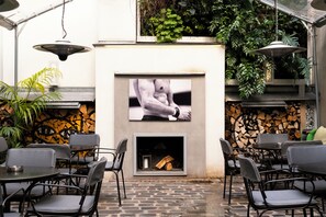 An outdoor seating area with tables and chairs, a fireplace, and a wall-mounted TV screen displaying an image. - Hotel Jules & Jim (Paris)