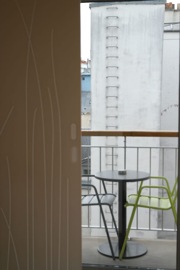 A balcony with a table and chairs, a view of a building, and a window with a curtain.