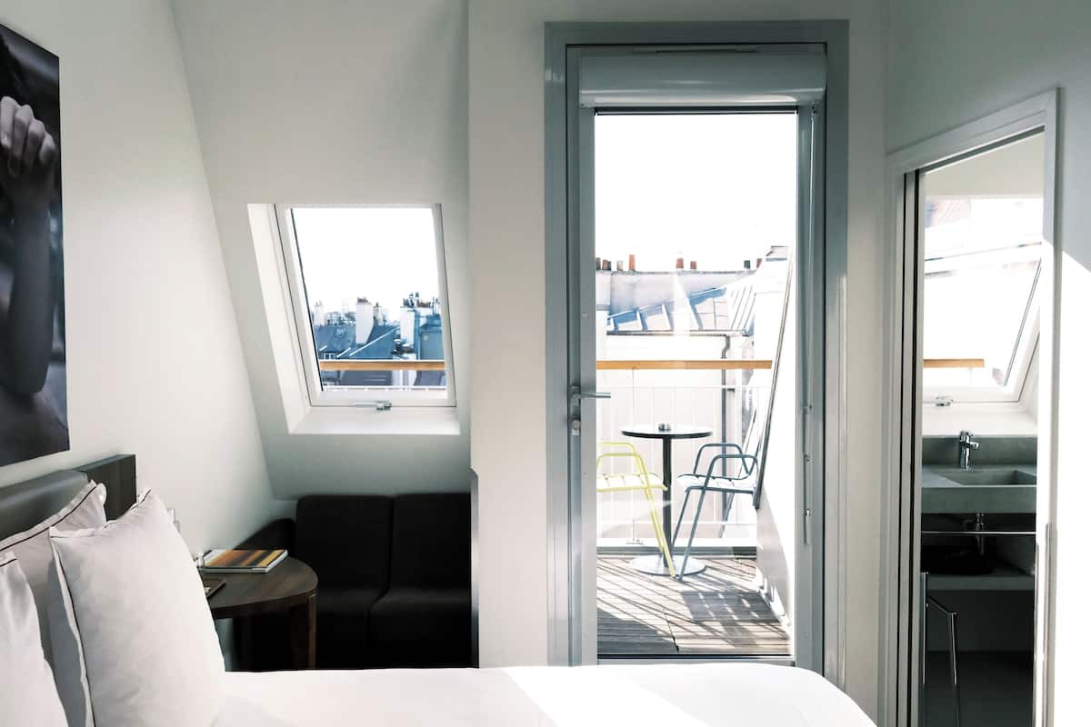 A hotel room with a bed, a small table with a book, a chair, a window with a view of the city, and a balcony with chairs and a table.