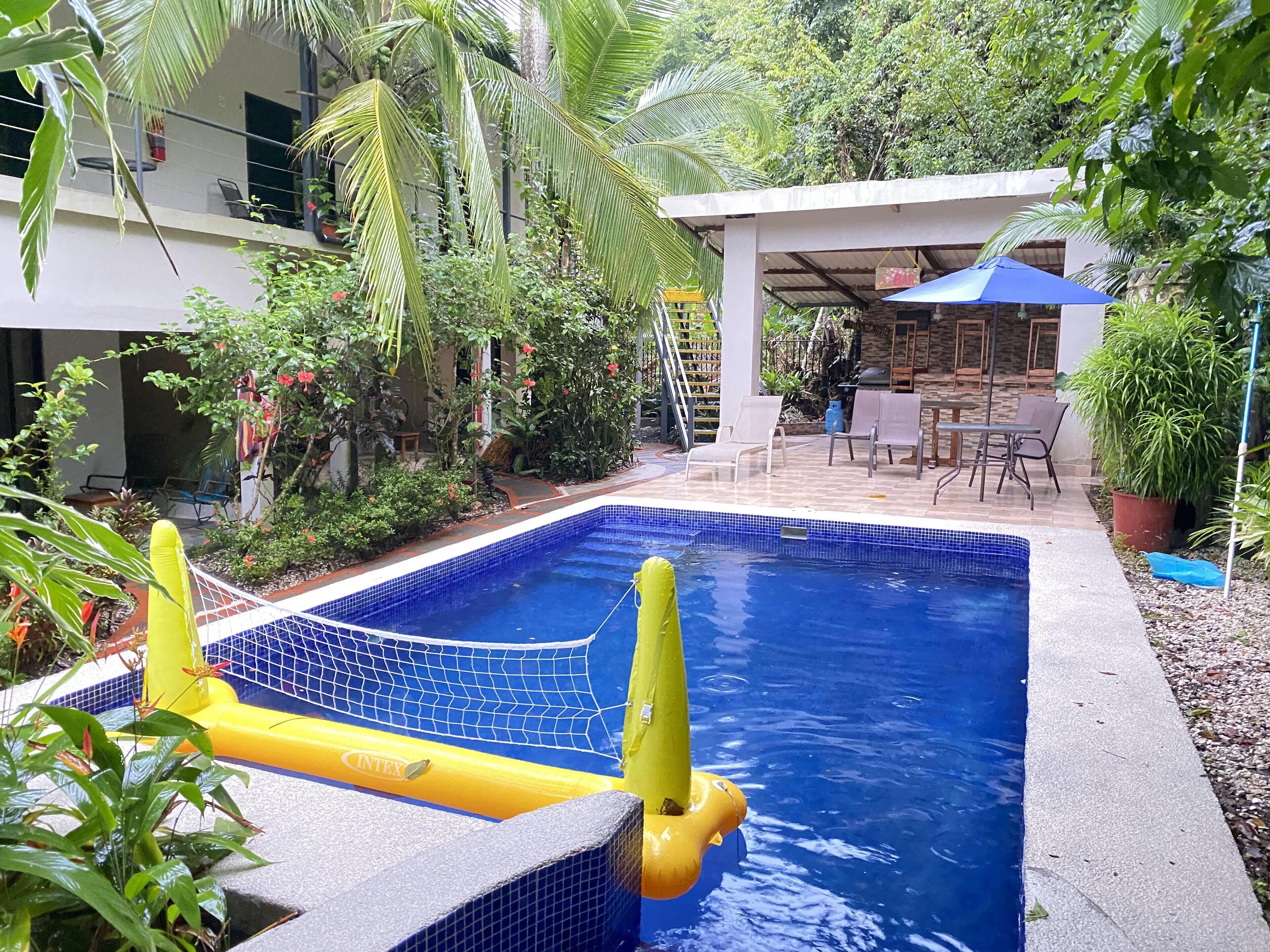 Una piscina con red de seguridad, rodeada de exuberante vegetación y una zona de estar cubierta.