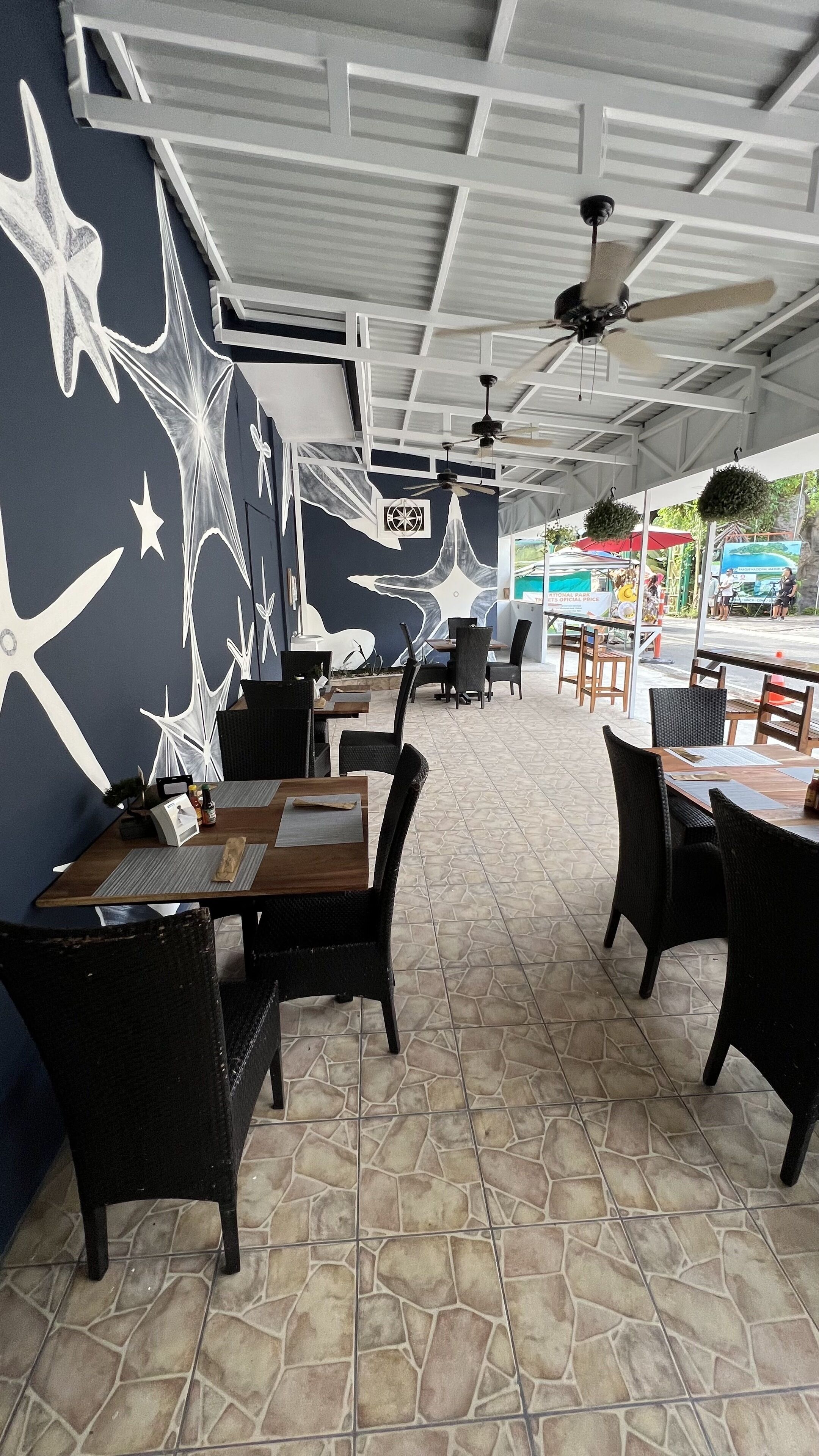 Interior de restaurante con arte mural en blanco y negro, piso de baldosas de piedra y sillas de mimbre negro.
