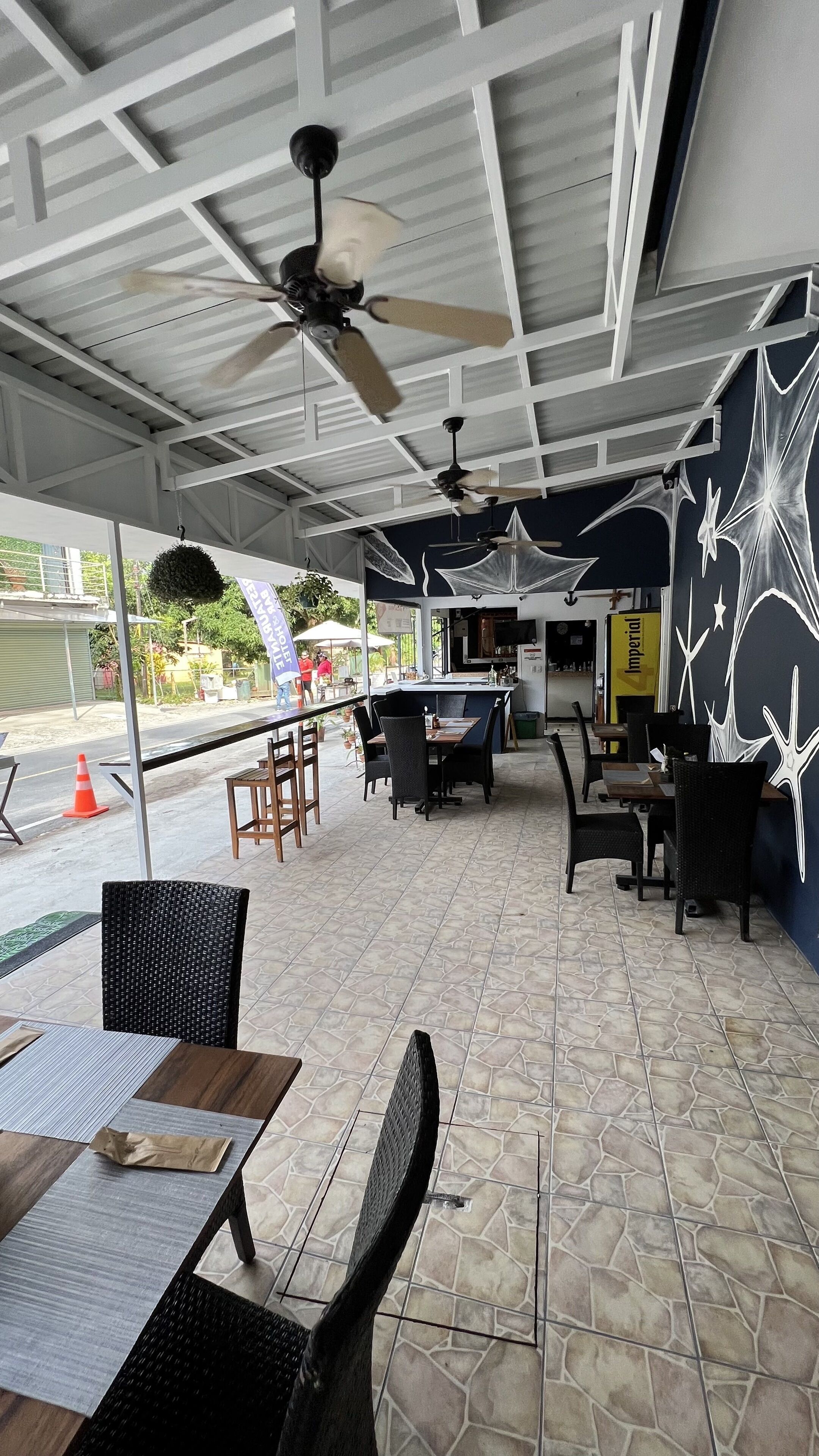 Un café en plein air avec des tables et des chaises, des ventilateurs de plafond et un sol carrelé.