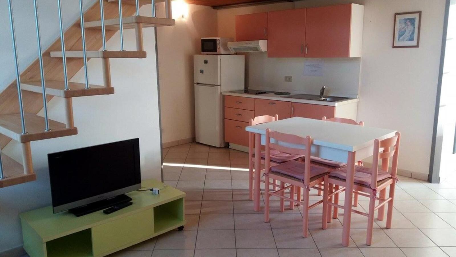 A compact living space with a TV on a green stand, a dining table with pink chairs, a kitchen area with a microwave and refrigerator, and a staircase with wooden steps.