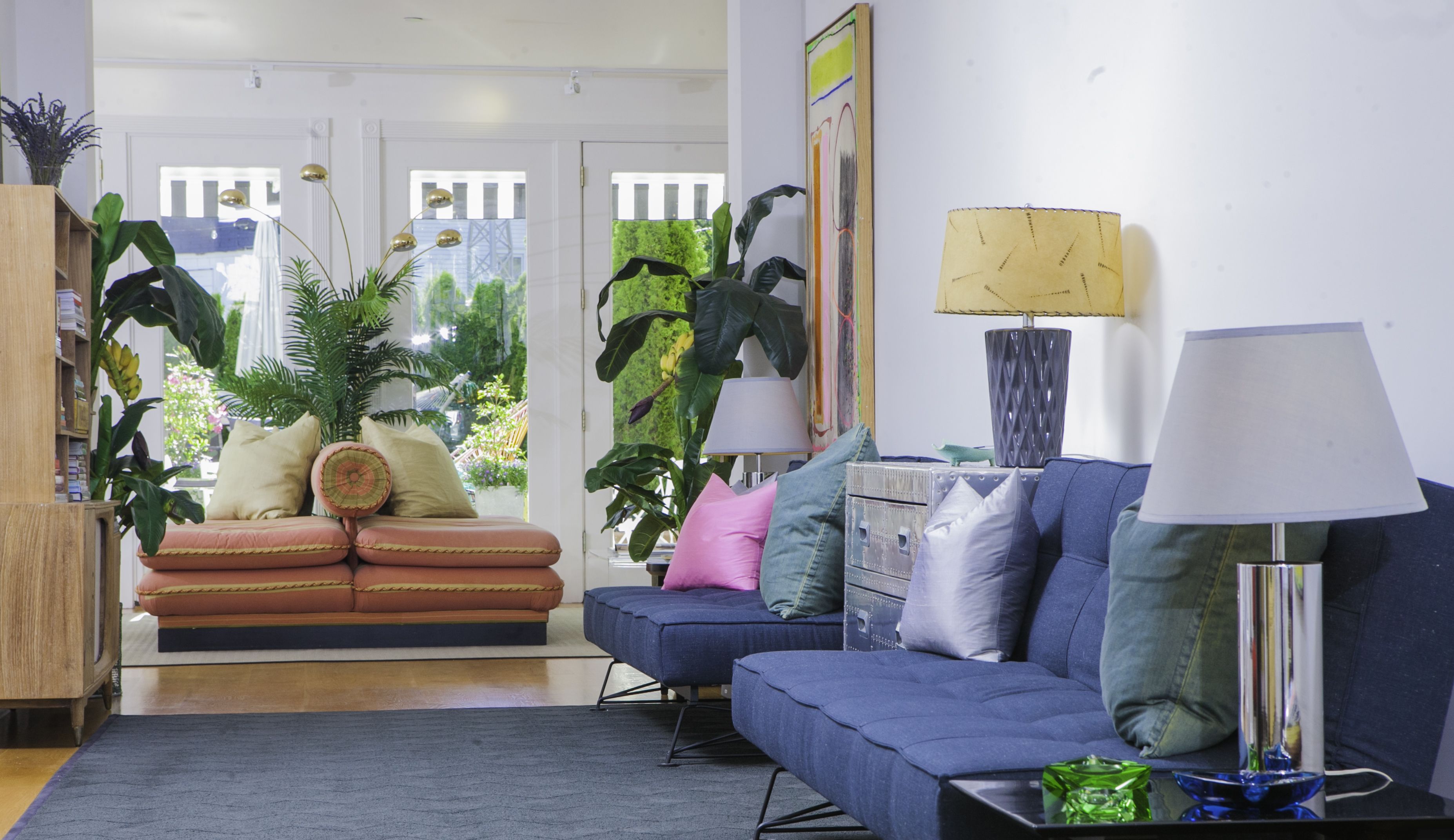 A living room with a sofa, a blue couch, a coffee table, and a lamp. There are plants and a painting on the wall.