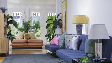 A living room with a sofa, a blue couch, a coffee table, and a lamp. There are plants and a painting on the wall.