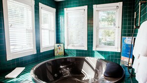 A bathroom with a bathtub, green tiled walls, and windows with white blinds. - Zey Hotel (Greenport)