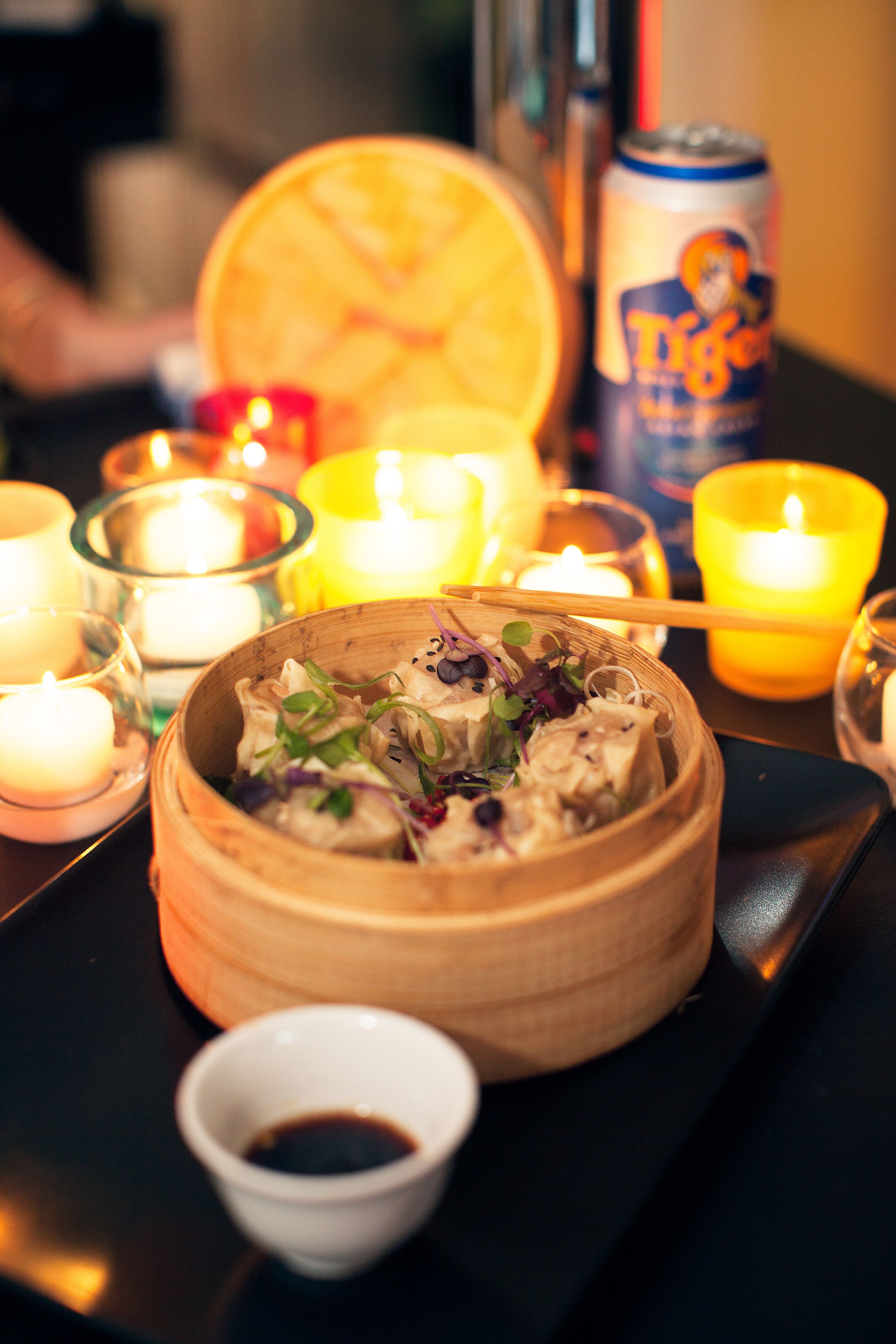 A dimly lit table with a bamboo steamer containing food, lit candles, and a can of Tiger beer.