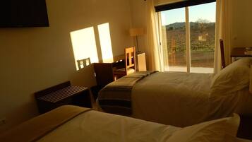 Quarto de hotel com duas camas, uma escrivaninha, uma cadeira, um abajur e uma janela grande com vista para o exterior.