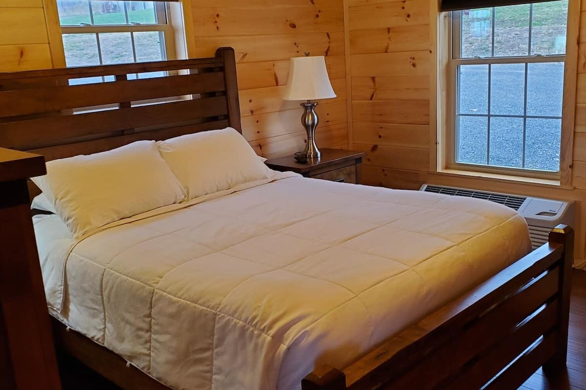 A wooden cabin bedroom with a bed, two windows, a nightstand with a lamp, and a wooden headboard.
