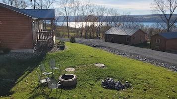 A grassy outdoor area with a fire pit, a bench, and a small pile of rocks. There are wooden buildings and a view of a lake in the background.