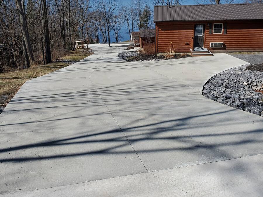 A wooden cabin with a concrete driveway leading to a dock by a lake.