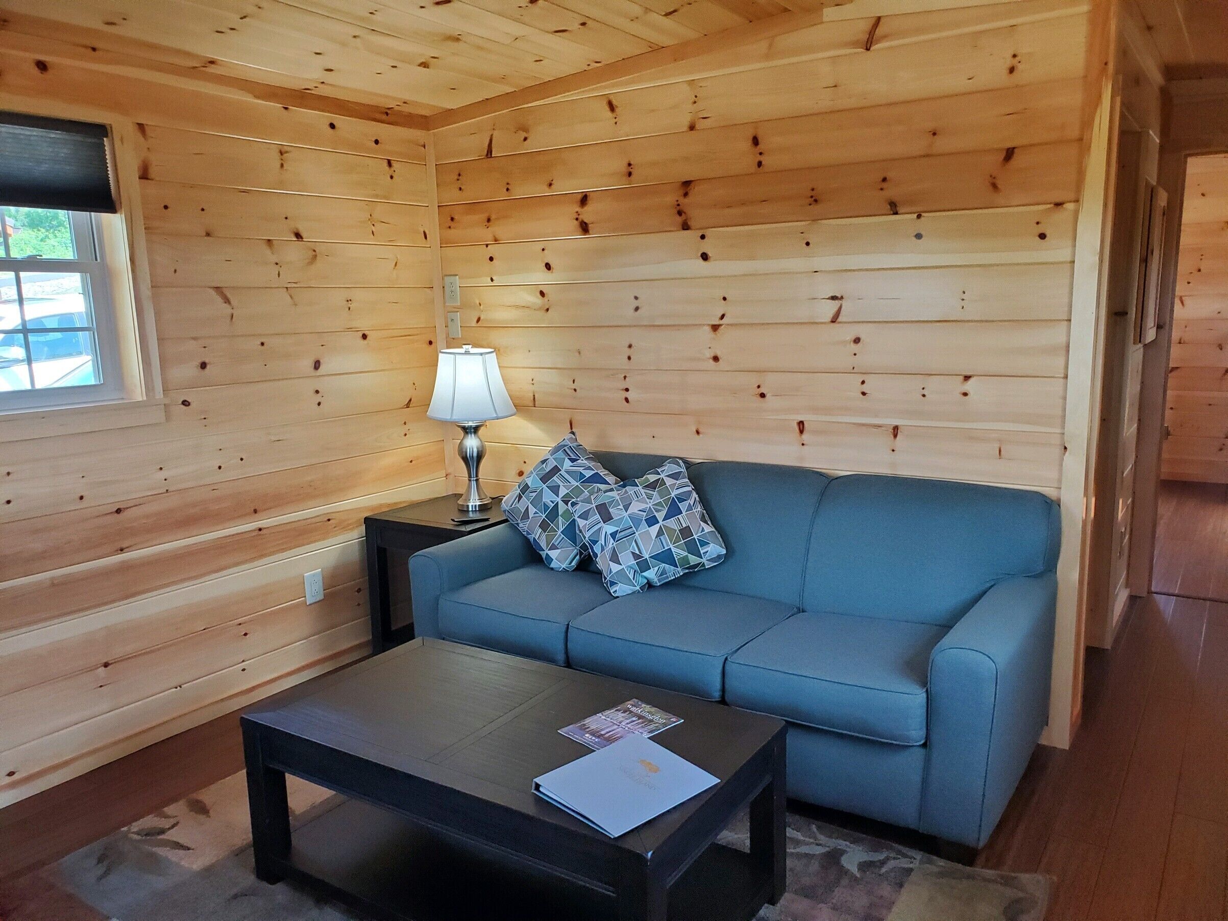 A cozy room with wooden walls, a blue sofa, a small table with a lamp, and a window.