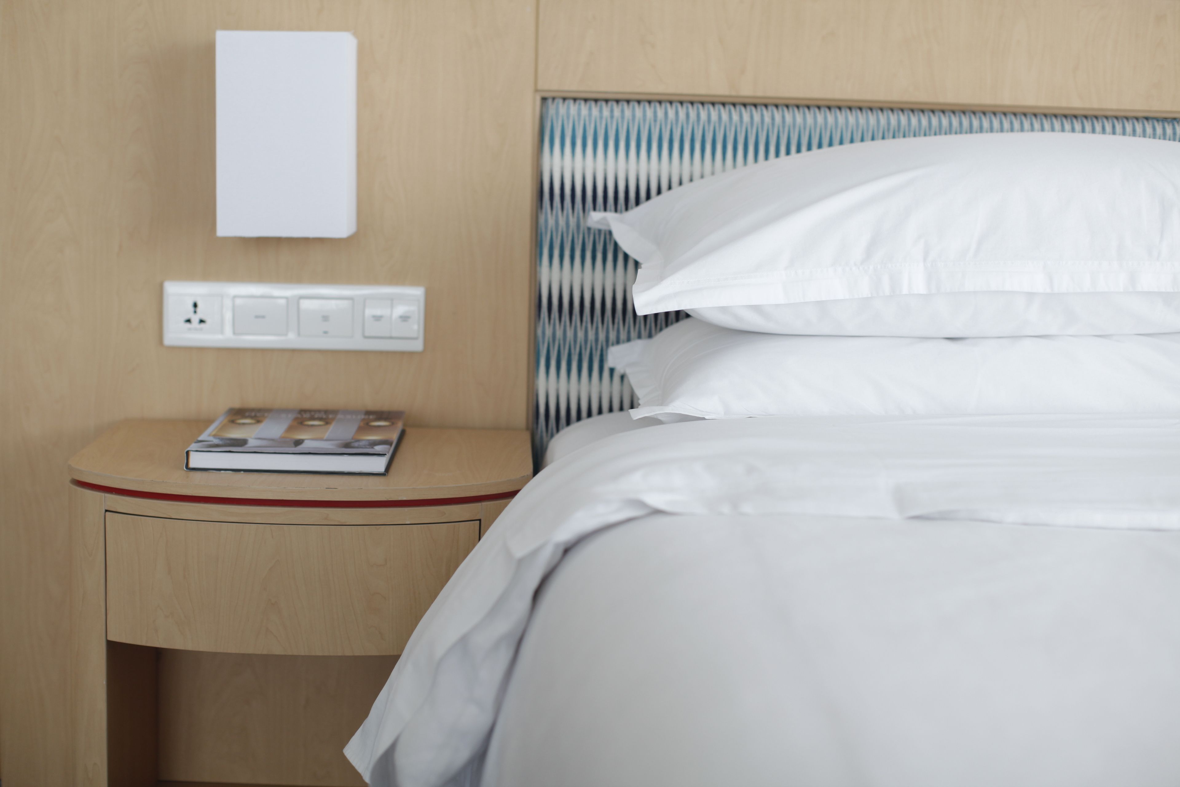 A neatly made bed with white linens, a bedside table with a book, and a wall-mounted light switch.