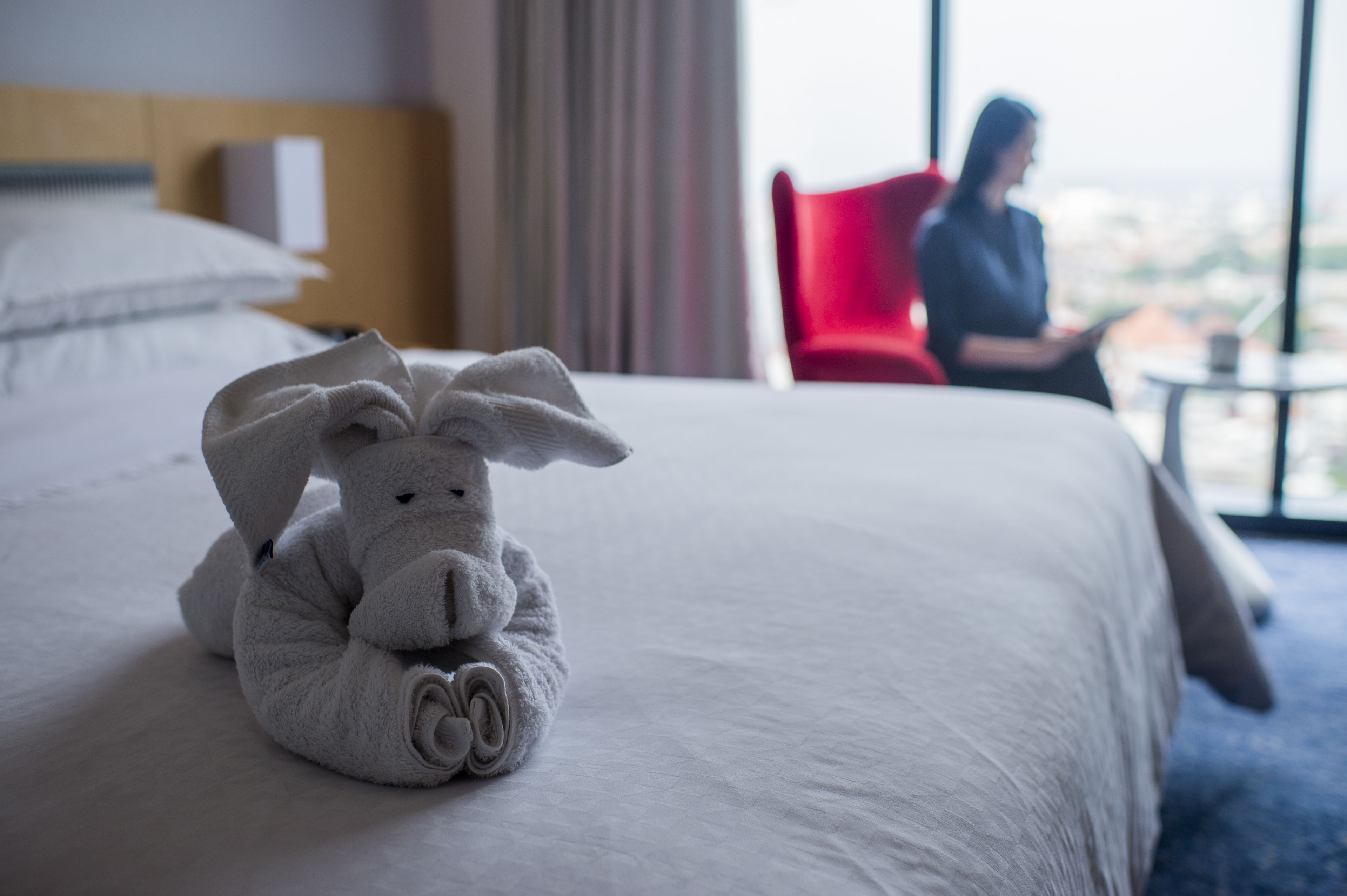 A neatly made bed with a rolled towel shaped like a bunny, a red chair, and a view of a cityscape through a window.
