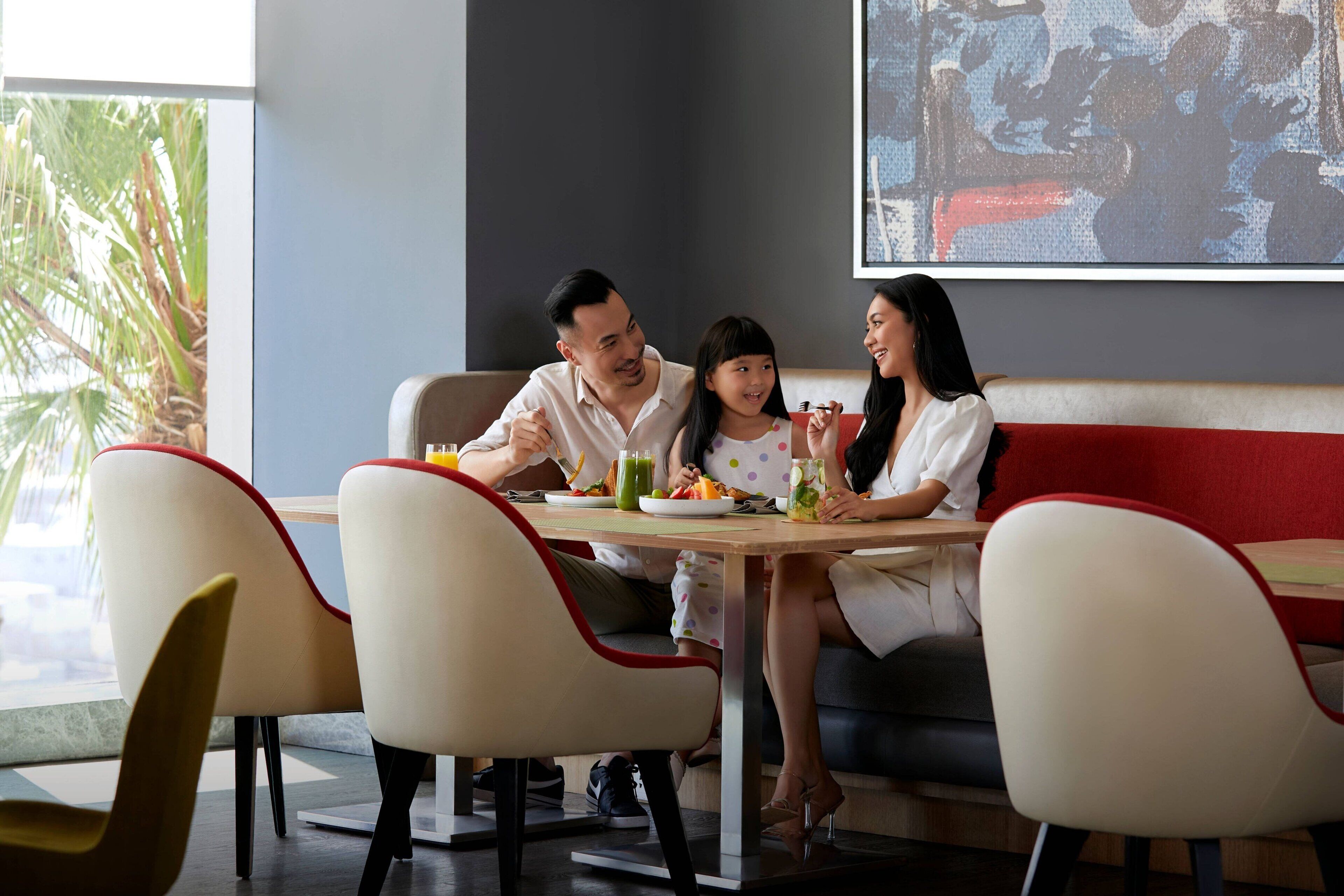 A family dining at a restaurant with a modern interior, including a large screen displaying a scene, a casual seating arrangement, and a view of palm trees outside.