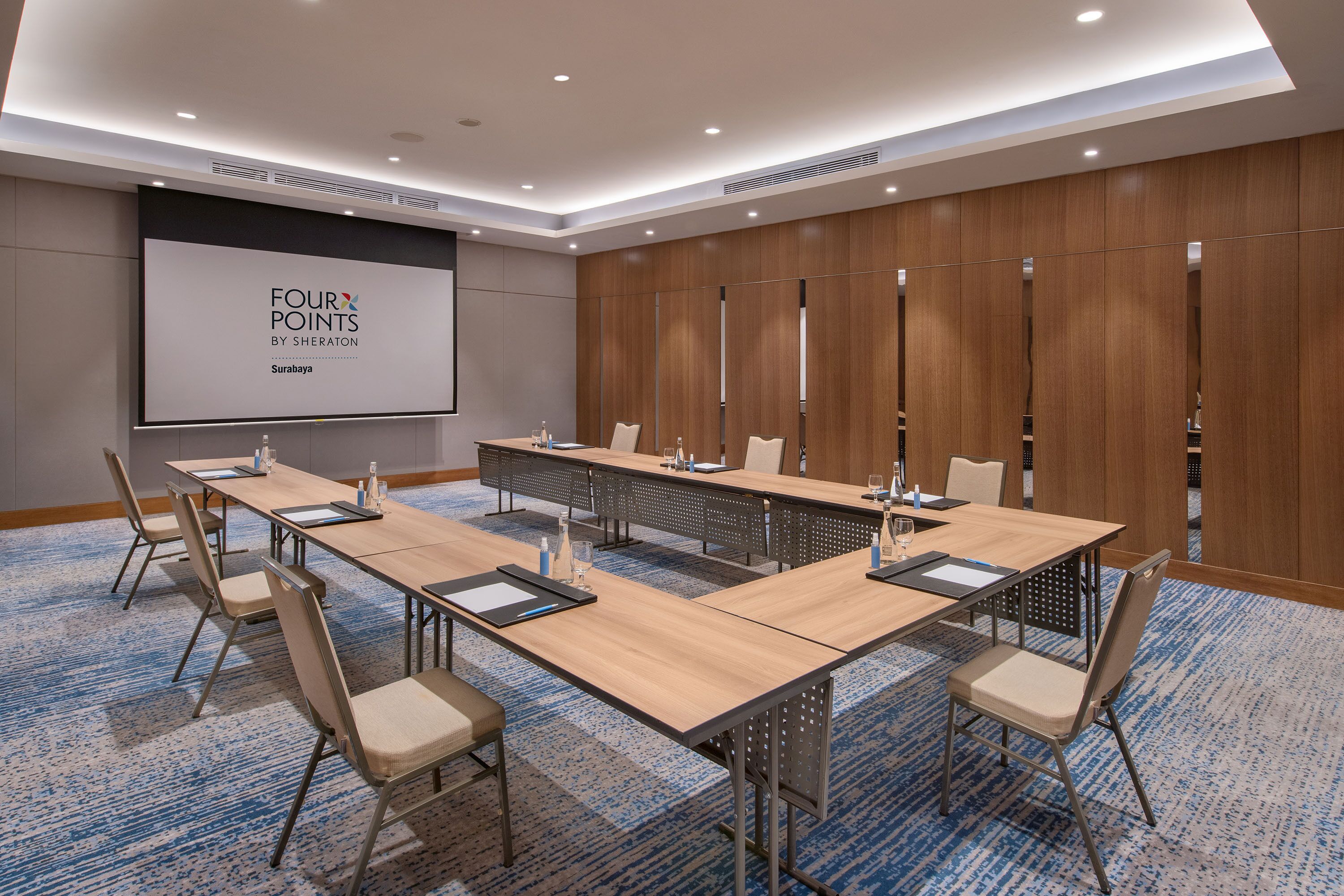 Une salle de conférence équipée d’une longue table, de chaises, d’un écran de projection et d’un mur recouvert de panneaux en bois.