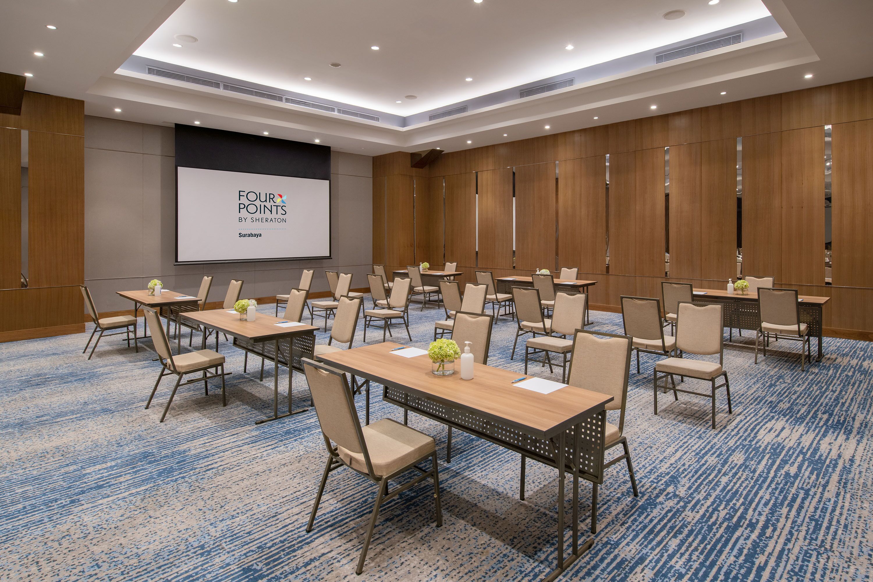 A conference room with multiple tables and chairs, a large screen, and a blue carpeted floor.