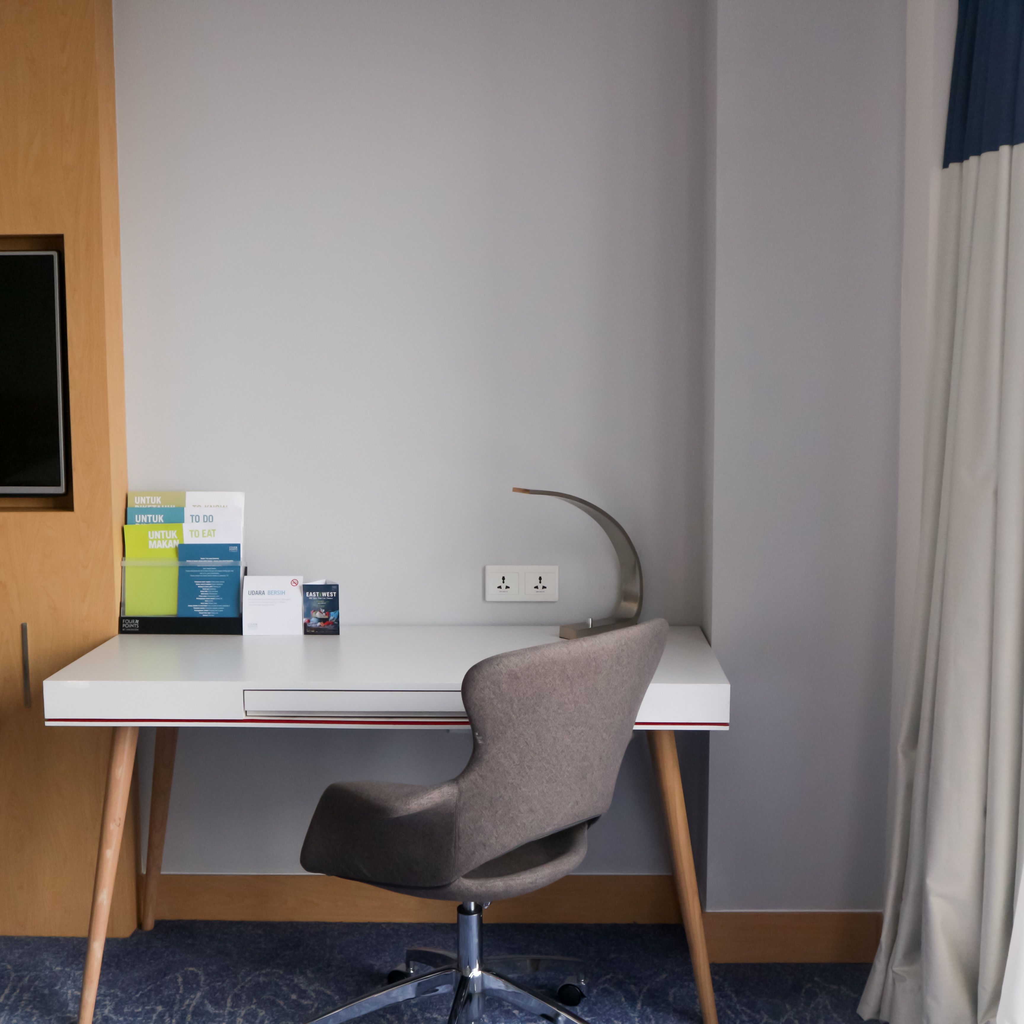 A modern hotel room with a white desk, a grey chair, and a small bookshelf with books and a magazine.